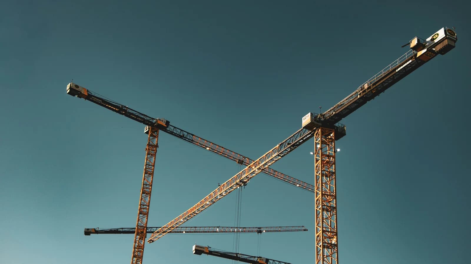 Grúas de construcción con estructuras oscuras y detalles dorados contra un cielo azul grisáceo